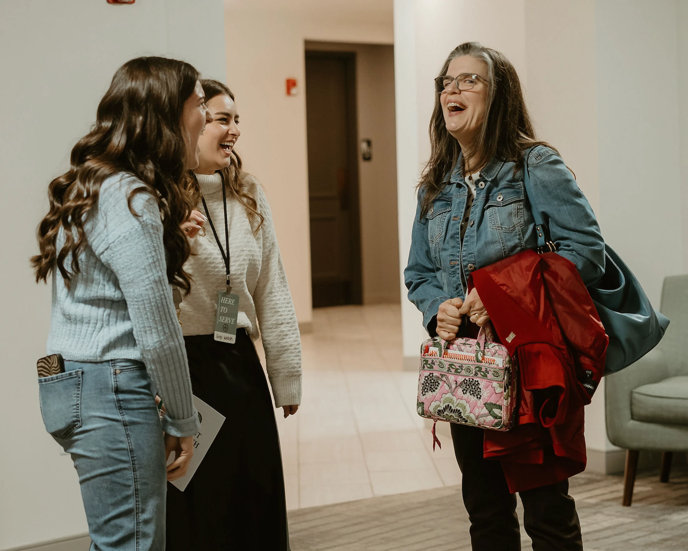 Three women of different ages stand in a brightly lit hallway, laughing together. One young woman wears a lanyard that says 'HERE TO SERVE'.