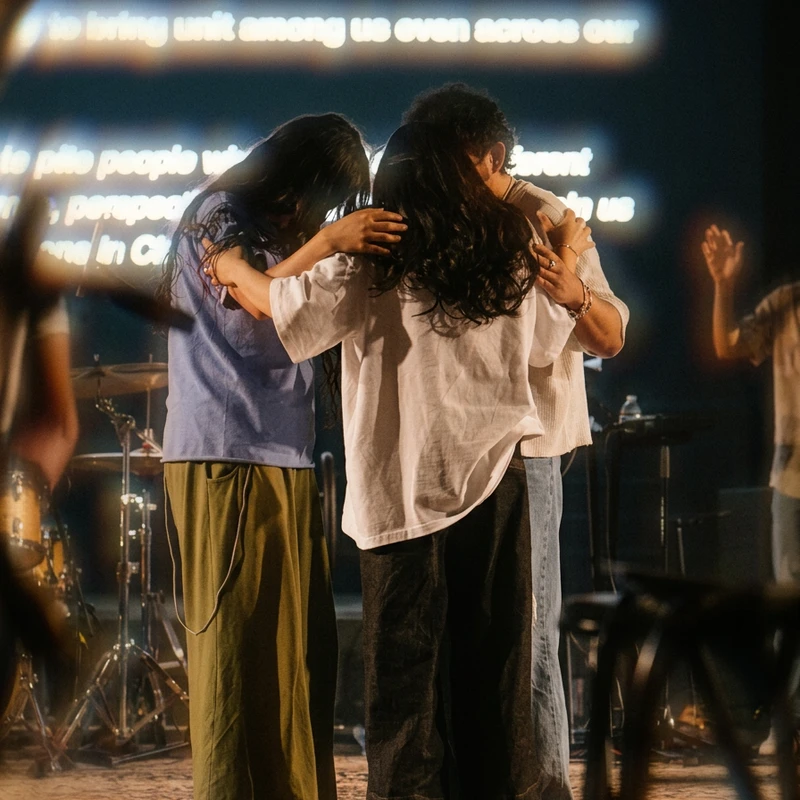 Three people stand on a stage in a group hug, their heads bowed together. Behind them, a screen displays glowing text and a drum kit is visible.