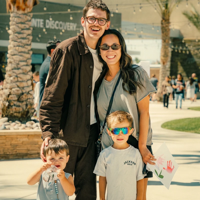 A smiling family of four poses for a photo outdoors. The parents stand behind their two young sons, one of whom holds a red handprint painting.