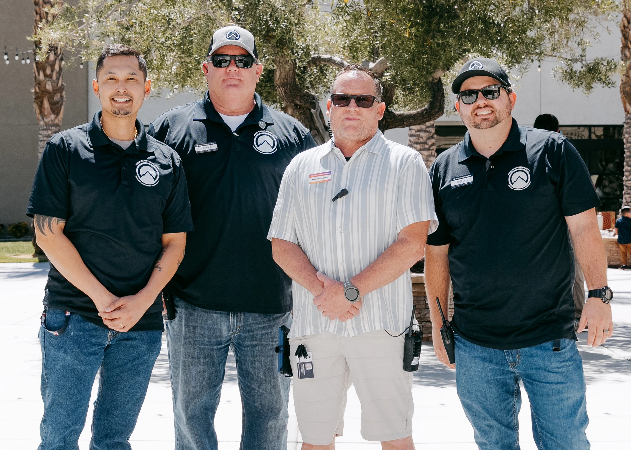 Four men from a campus safety team stand together for a group photo outdoors. Three wear black polo shirts with logos, and one wears a striped button-up shirt.