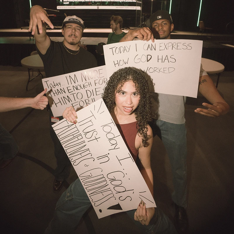 A group of three diverse young people hold up handwritten signs with messages about their Christian faith, posing for a photo in a large room.