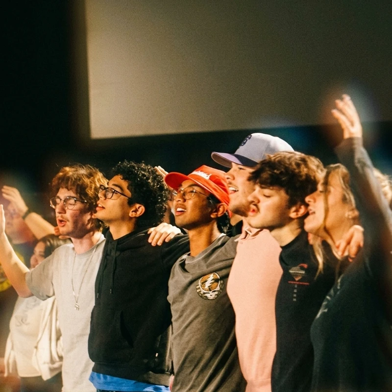 A diverse group of young people stand with their arms around each other, looking up and singing with passionate expressions in a dimly lit setting.