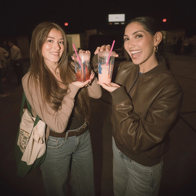 Two young women smile at the camera, holding up their colorful Dutch Bros drinks with pink straws in a dimly lit indoor space.