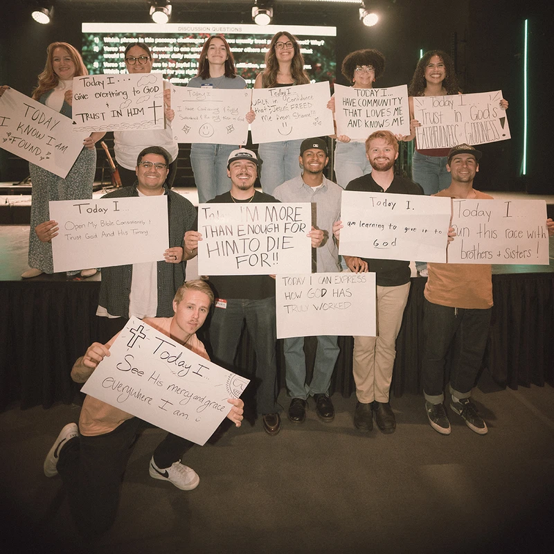 A diverse group of young adults stands on a stage, smiling and holding up handwritten signs sharing their personal affirmations of faith and trust in God.