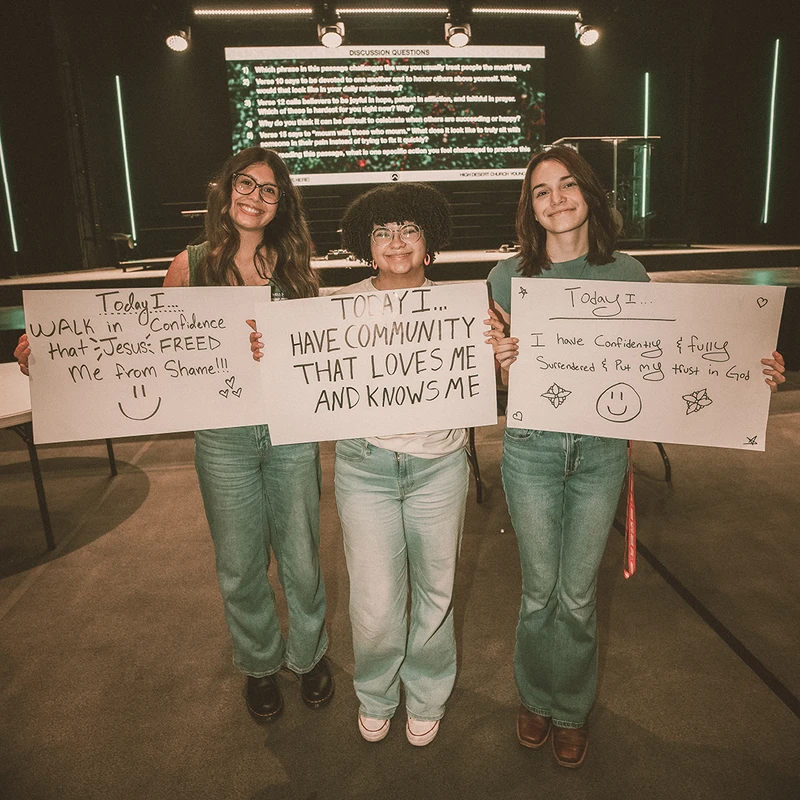 Three young women in a church auditorium smile while holding signs with handwritten messages about confidence, community, and trusting in God.
