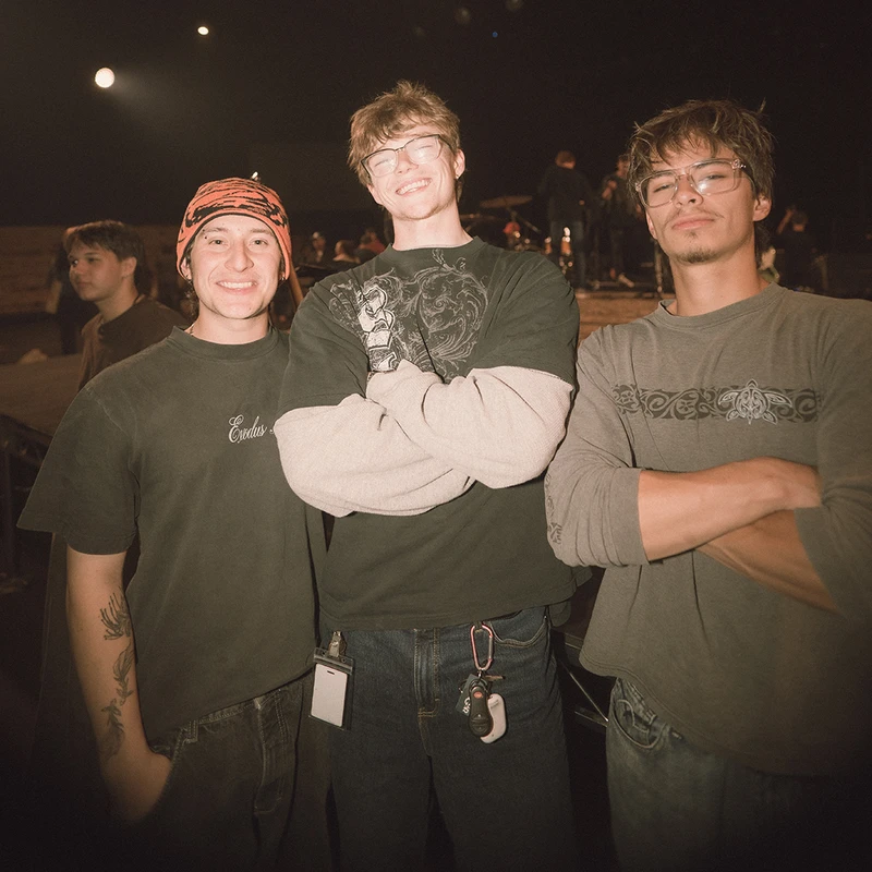 A grainy, warm-toned photo of three young men posing at a concert. Two have their arms crossed, and they all look towards the camera, smiling.