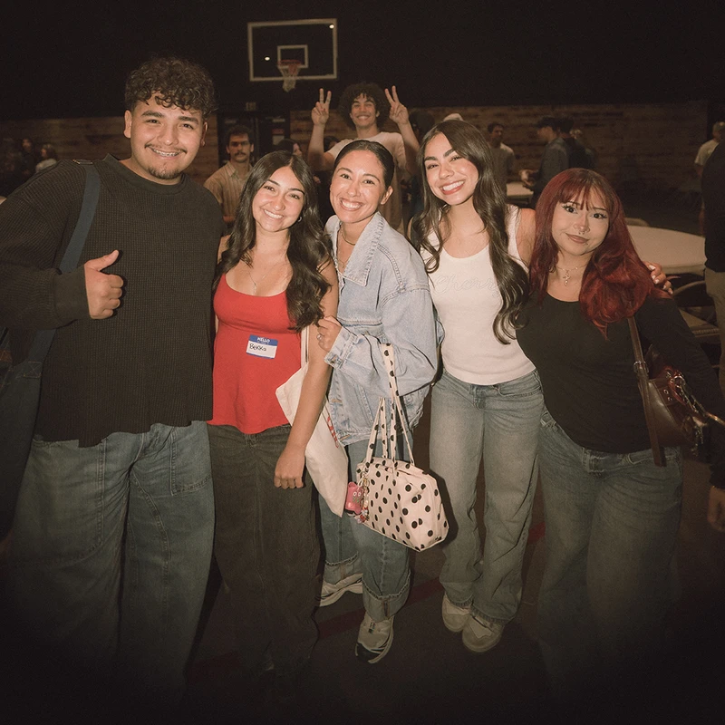 A group of six diverse young friends smile and pose for a picture at an indoor event. A basketball hoop is visible in the background.