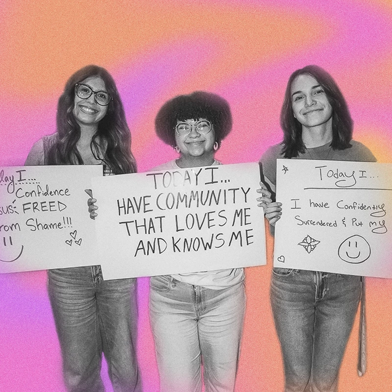 Three young women stand smiling, holding signs with handwritten positive affirmations against a pink and orange background. The photo is in black and white.