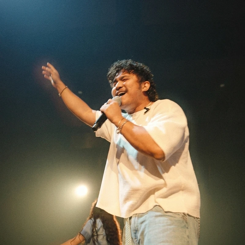 A man with curly hair and a white shirt sings passionately into a microphone on a dimly lit stage, with one arm raised in the air.