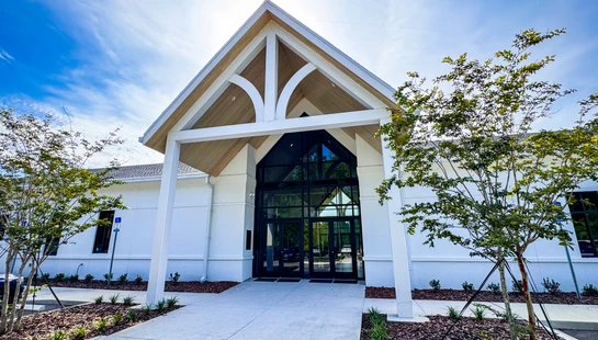 A low-angle shot of a modern white building's entrance, featuring a high gabled roof with exposed beams and a large glass entryway under a bright blue sky.