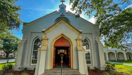 The front of a gray church with an open red door, seen from below on a bright, sunny day. The sun peeks from behind the cross on the roof against a blue sky.
