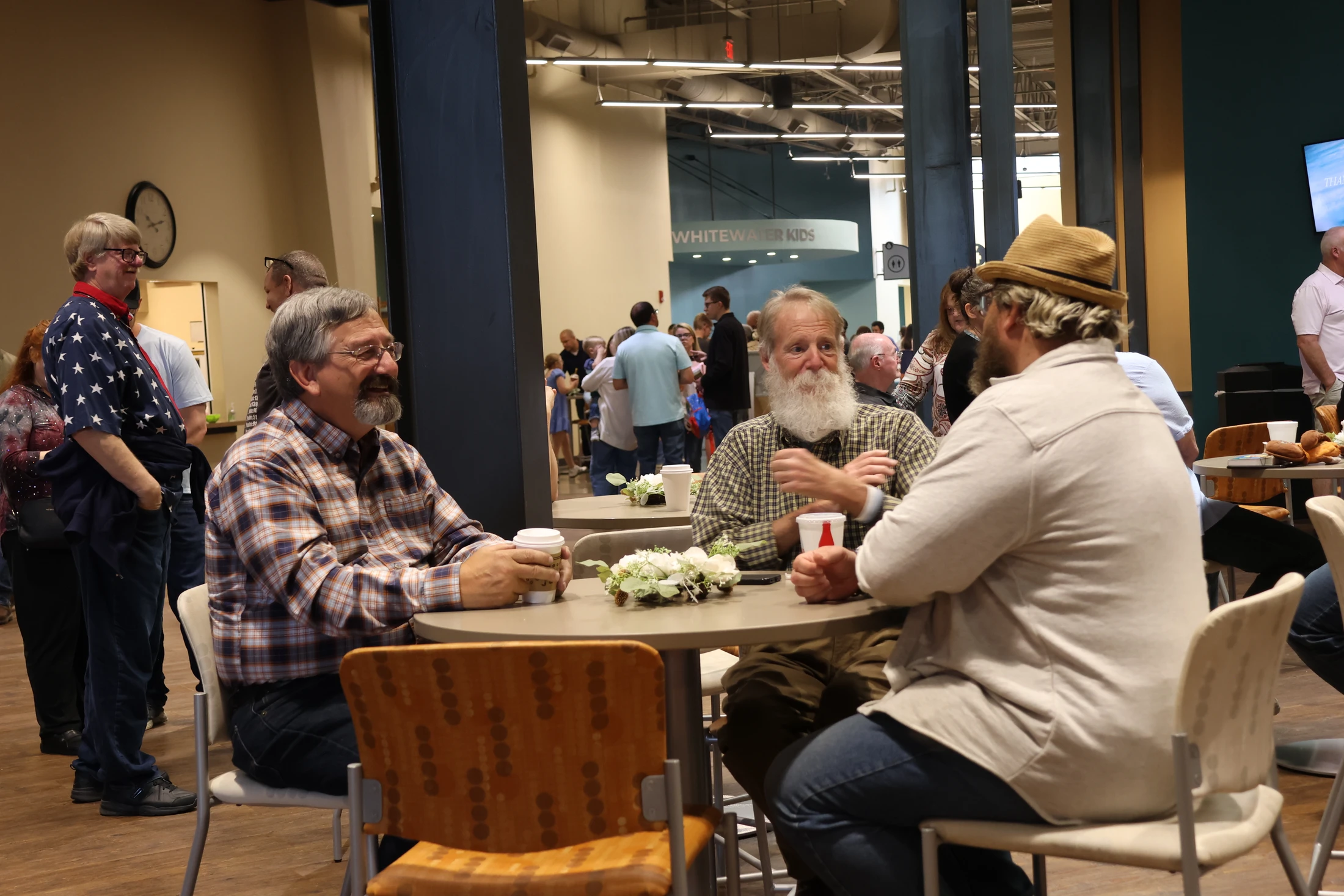 Three men sit at a round table having a conversation over coffee in a busy hall. One man has a long white beard, and another wears a straw hat.