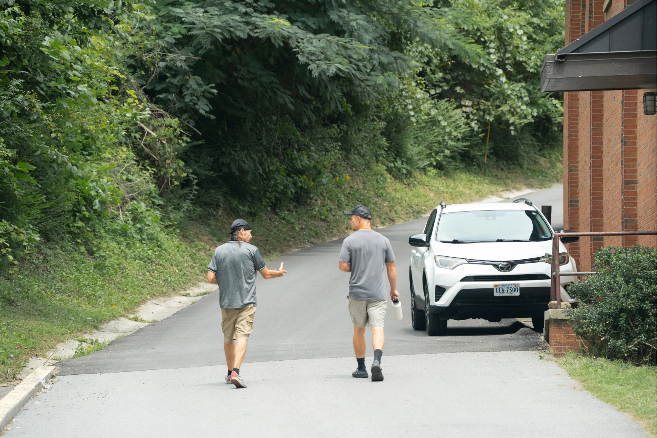 Two men in shorts and t-shirts walk up a paved road next to a wooded area. One man gestures as they talk. A white SUV is parked beside a brick building.