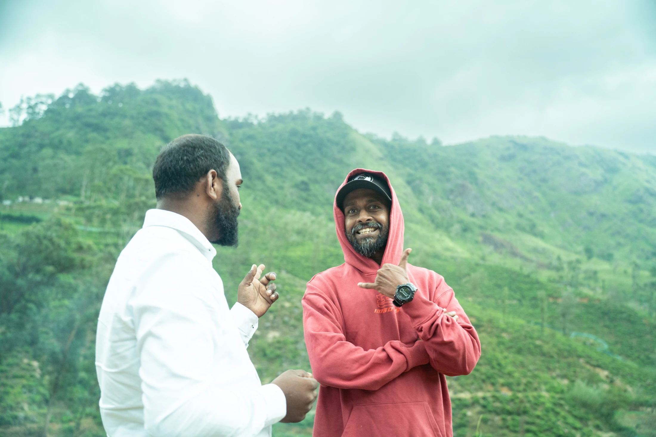 Two men stand in front of a vast green mountain landscape. One man in a red hoodie smiles at the camera, making a 'hang loose' hand gesture.
