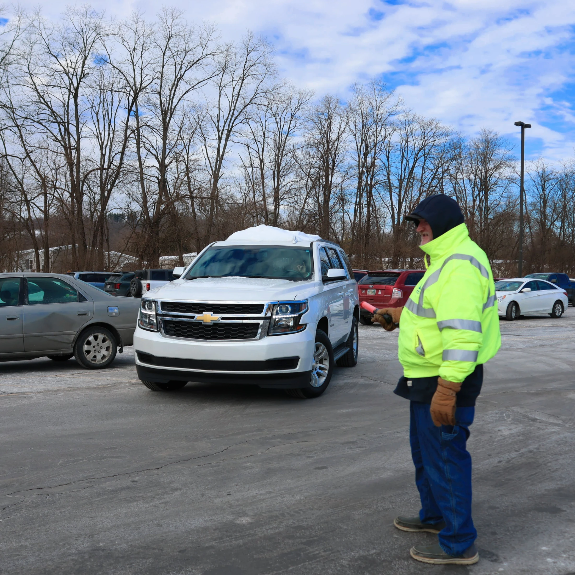 A man in a bright yellow safety jacket stands in a snowy parking lot, directing a white SUV with a red light wand on a cold winter day.