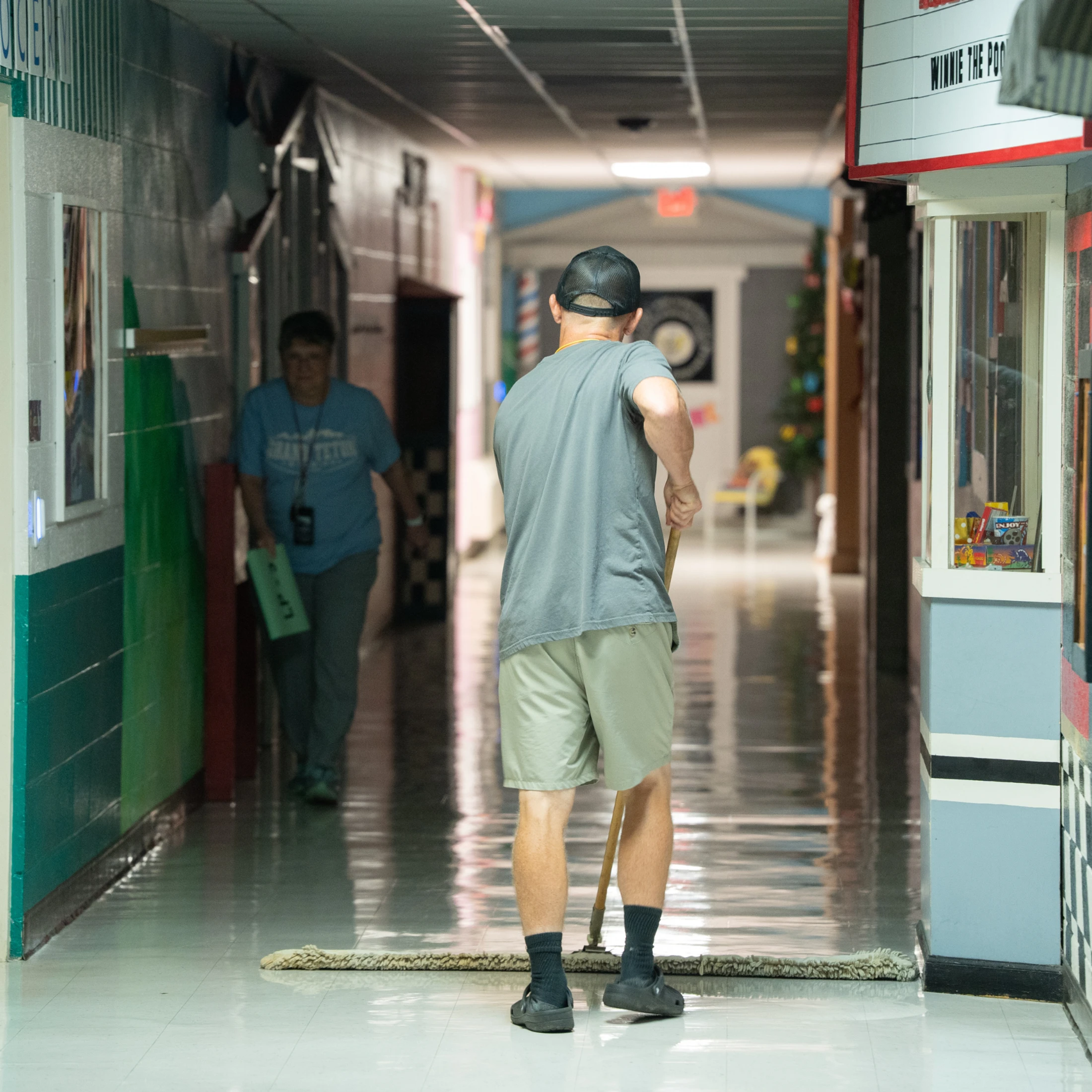 A man seen from behind uses a large dust mop to clean a long, shiny hallway floor. In the background, a woman walks down the hall.