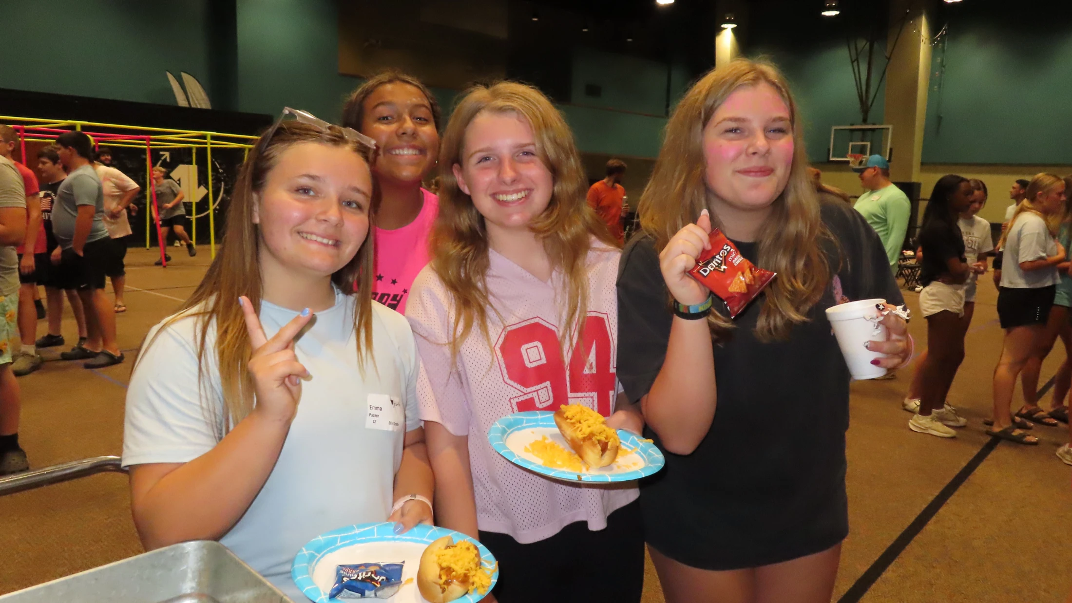 Four smiling teenage girls pose for a photo in a large room. They are holding paper plates with cheesy hot dogs and a bag of Doritos.