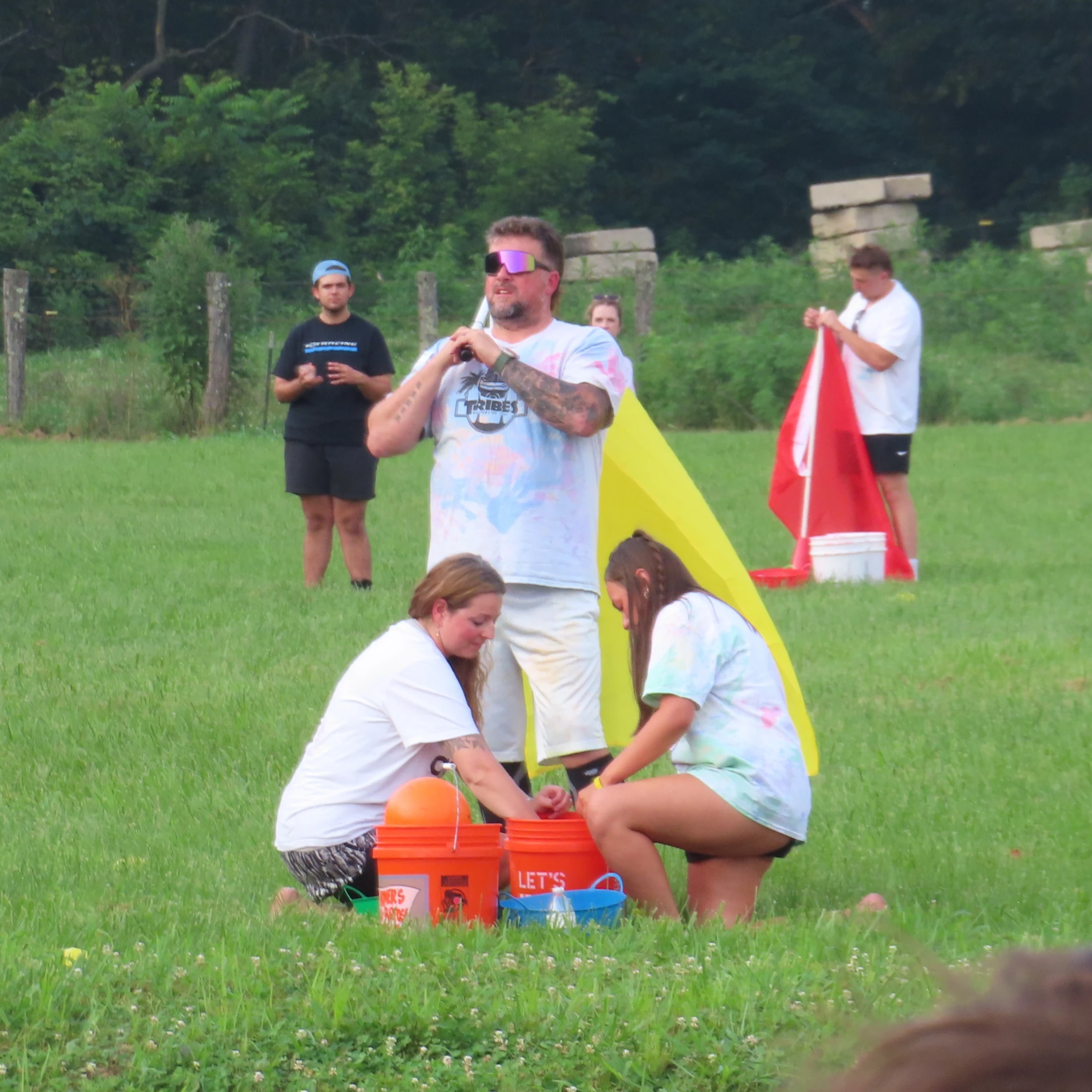 A group of people prepares for an outdoor game in a grassy field. Two women kneel by orange buckets while others stand and watch in the background.