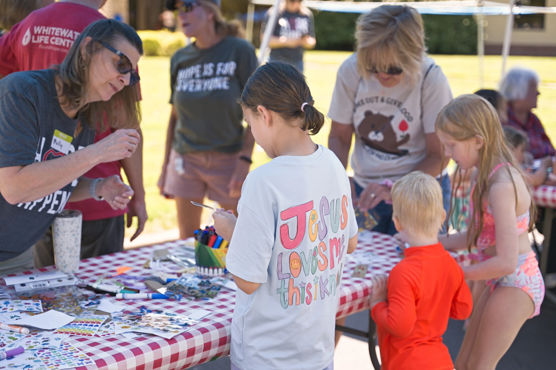 Children and adults gather around a red and white checkered table full of stickers and craft supplies during a sunny outdoor community event.
