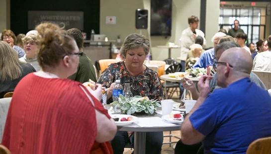 A group of adults eat and talk together while sitting around a small, round table in a large, crowded room during a community event.