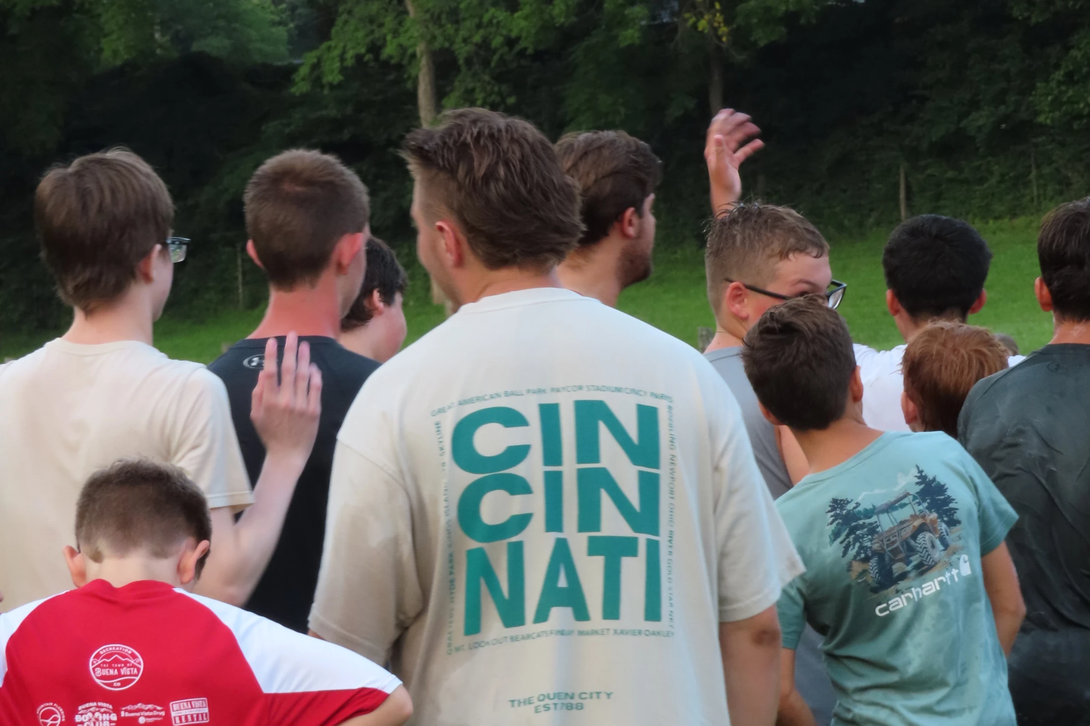 A view from behind a crowd of young men standing outdoors. One person wears a white t-shirt with "CINCINNATI" printed in large green block letters.