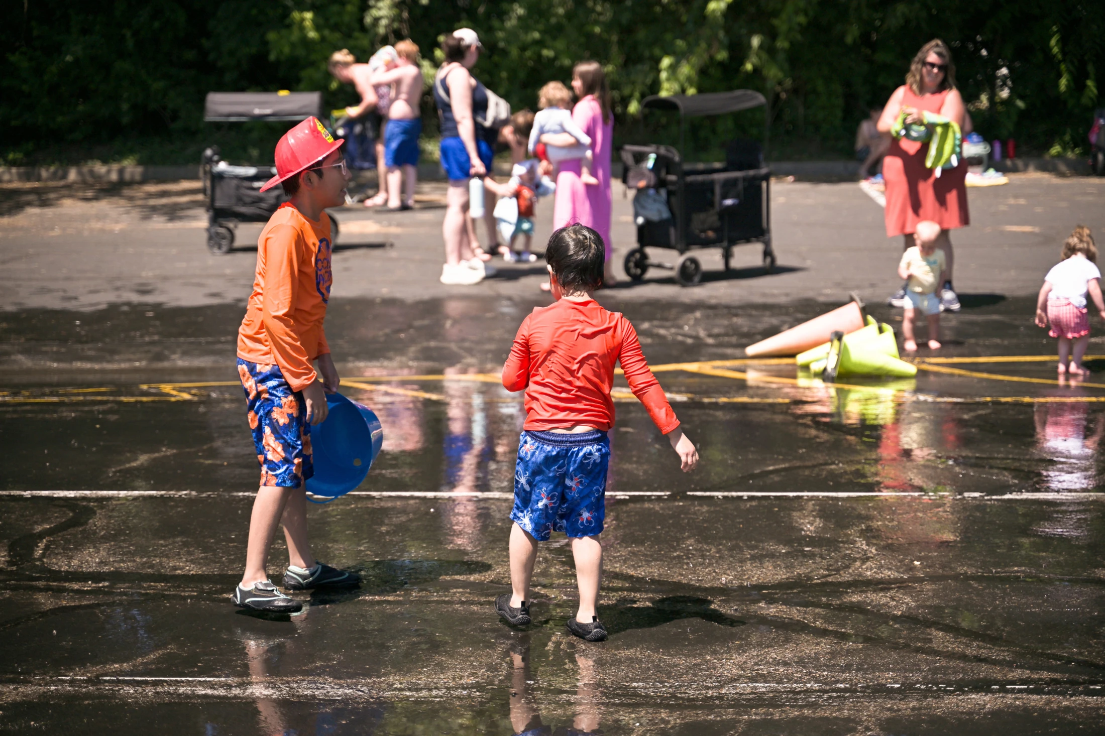 Two young boys in swimsuits play in a wet parking lot on a sunny day. One wears a red toy helmet and holds a blue bucket, with other families in the background.