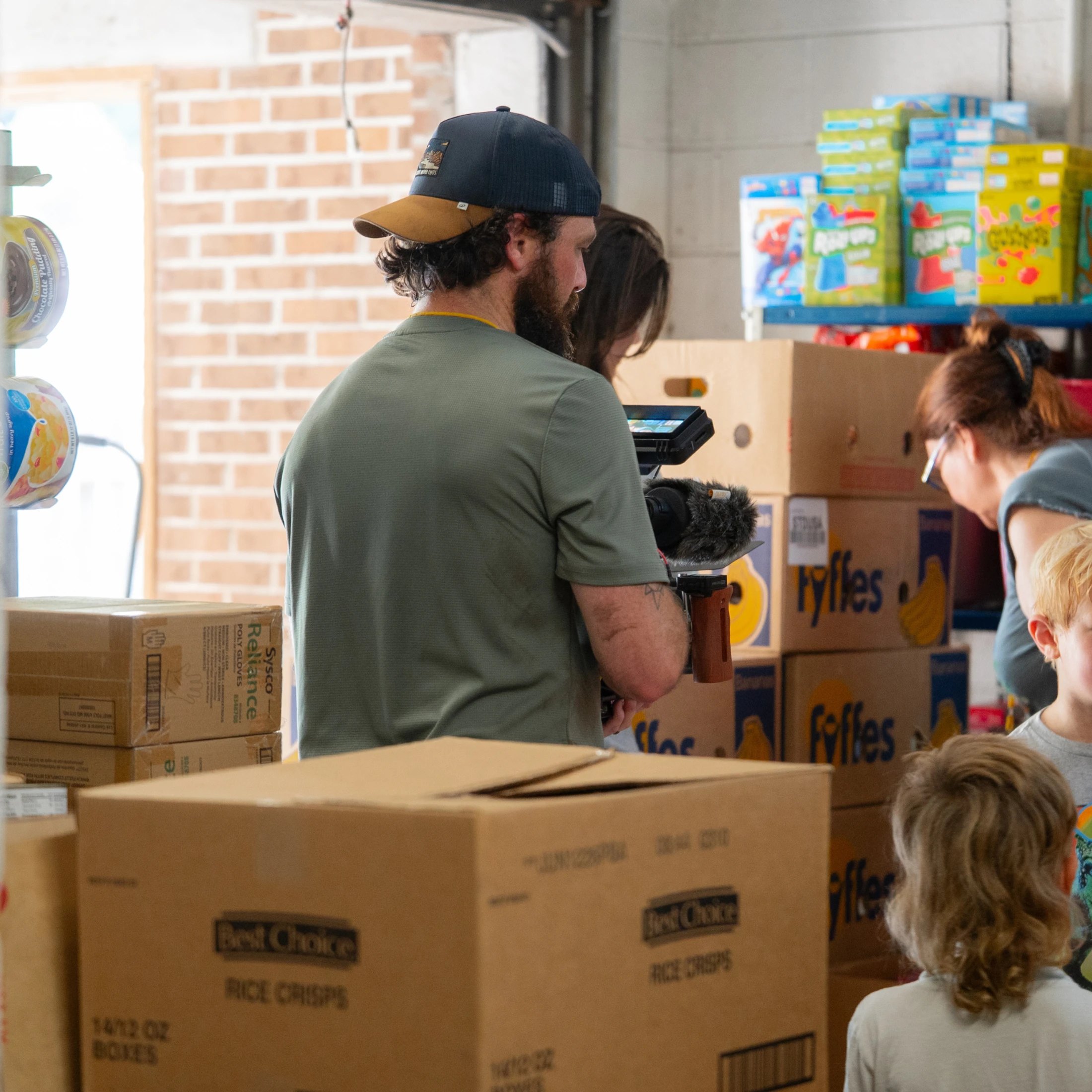 A man with a beard and a baseball cap holds a video camera, filming inside a warehouse-like room filled with cardboard boxes of food and other people.