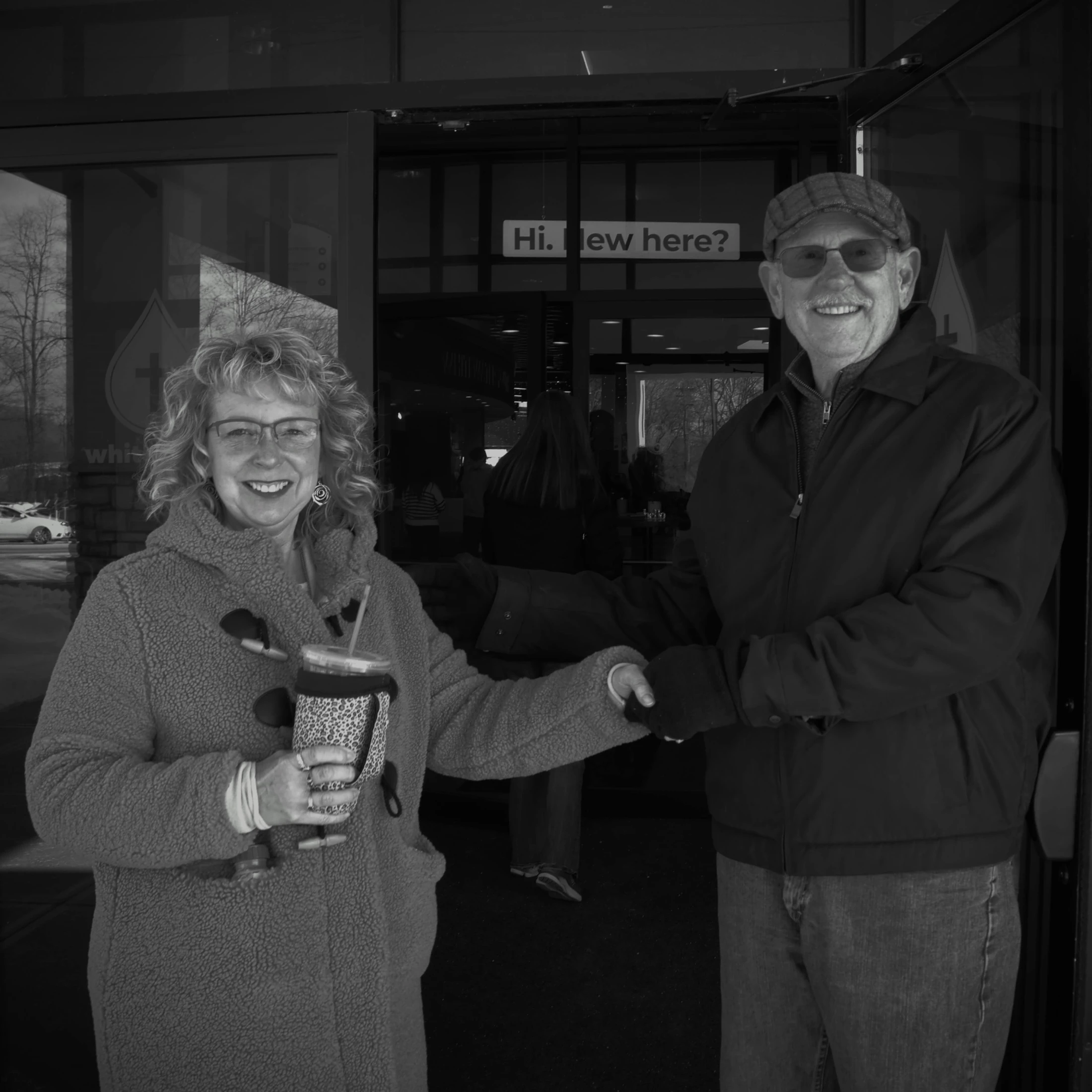 A black and white photo of a smiling man and woman shaking hands in front of a glass door. A sign in the background reads, 'Hi. New here?'.