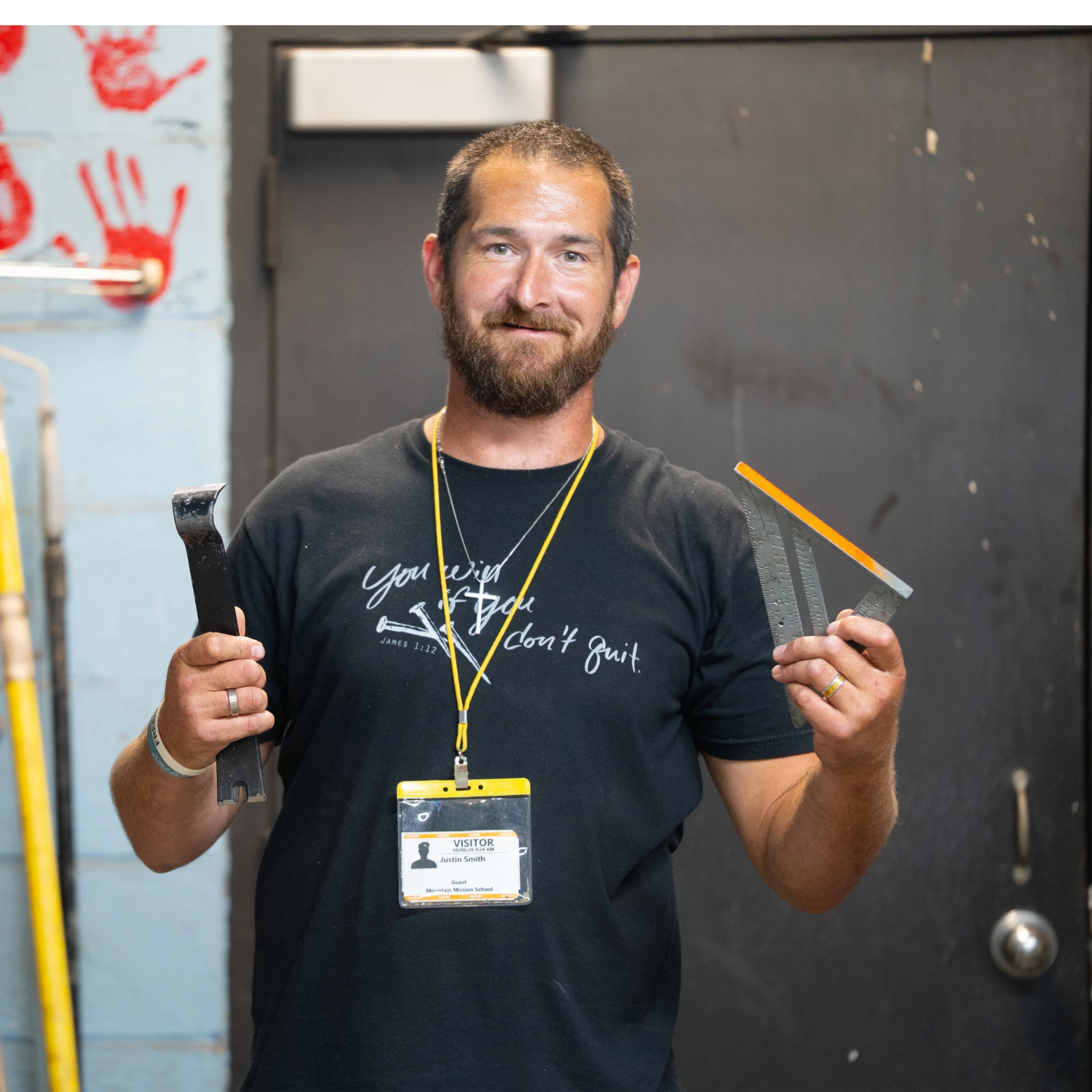A man with a beard wearing a black t-shirt and a visitor badge smiles while holding a pry bar in one hand and a speed square in the other.