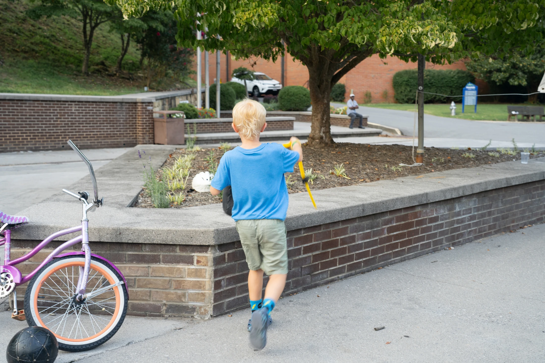 A young, blond-haired boy in a blue shirt runs away from the camera on a sidewalk, with a purple children's bicycle parked to the left in a park setting.