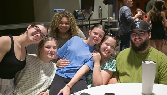 A group of six young adults smile and pose together for a photo while sitting around a white table at an indoor gathering.