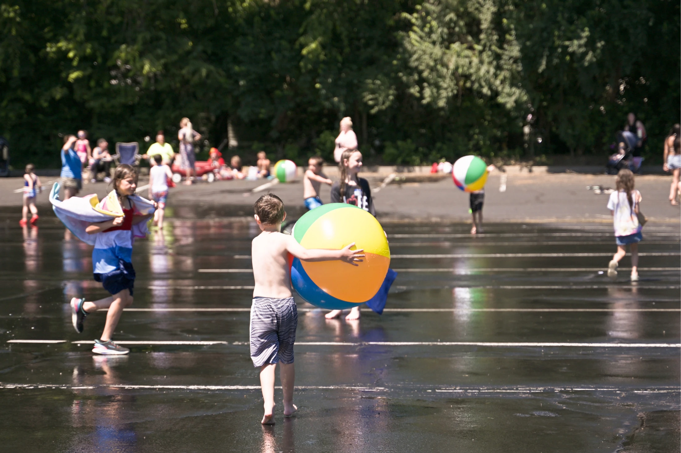 A shirtless young boy holds a large colorful beach ball while walking on a wet parking lot. Other children play in the water around him on a sunny day.