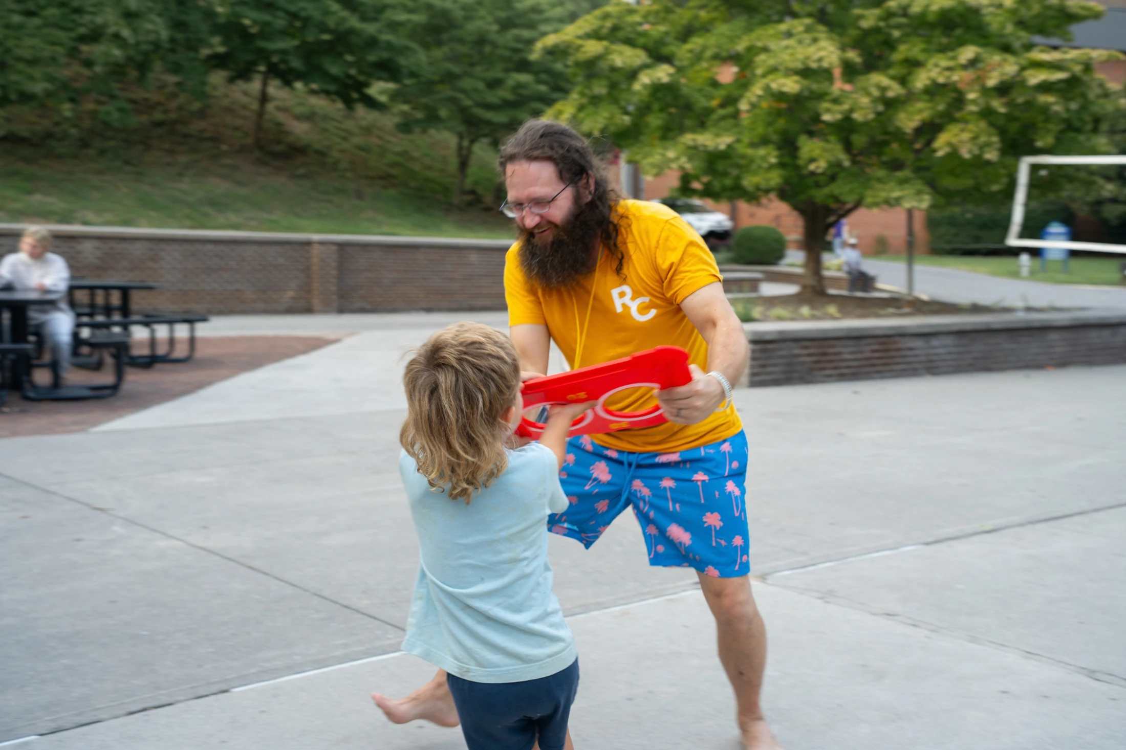 A smiling man with a long beard and a young child playfully tug on a large red plastic toy. They are standing on a concrete patio with trees in the background.