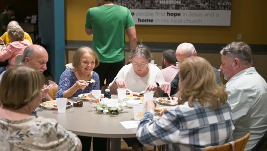 A group of older adults are gathered around a circular table, smiling and eating a meal together in what appears to be a church hall.