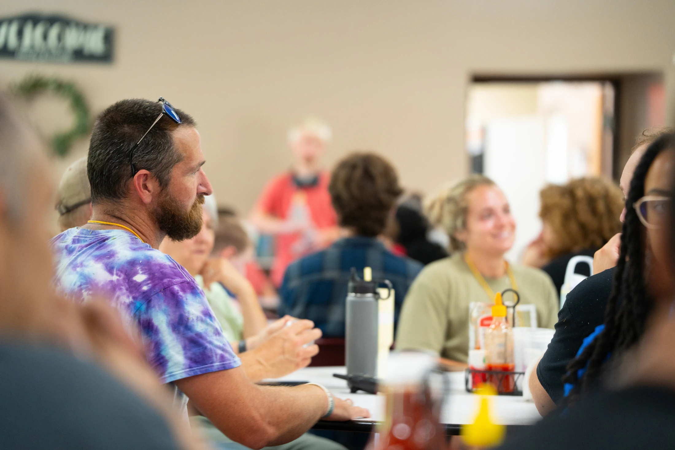 A man with a beard, wearing a purple tie-dye shirt and sunglasses on his head, sits at a table in a crowded room during a casual event.