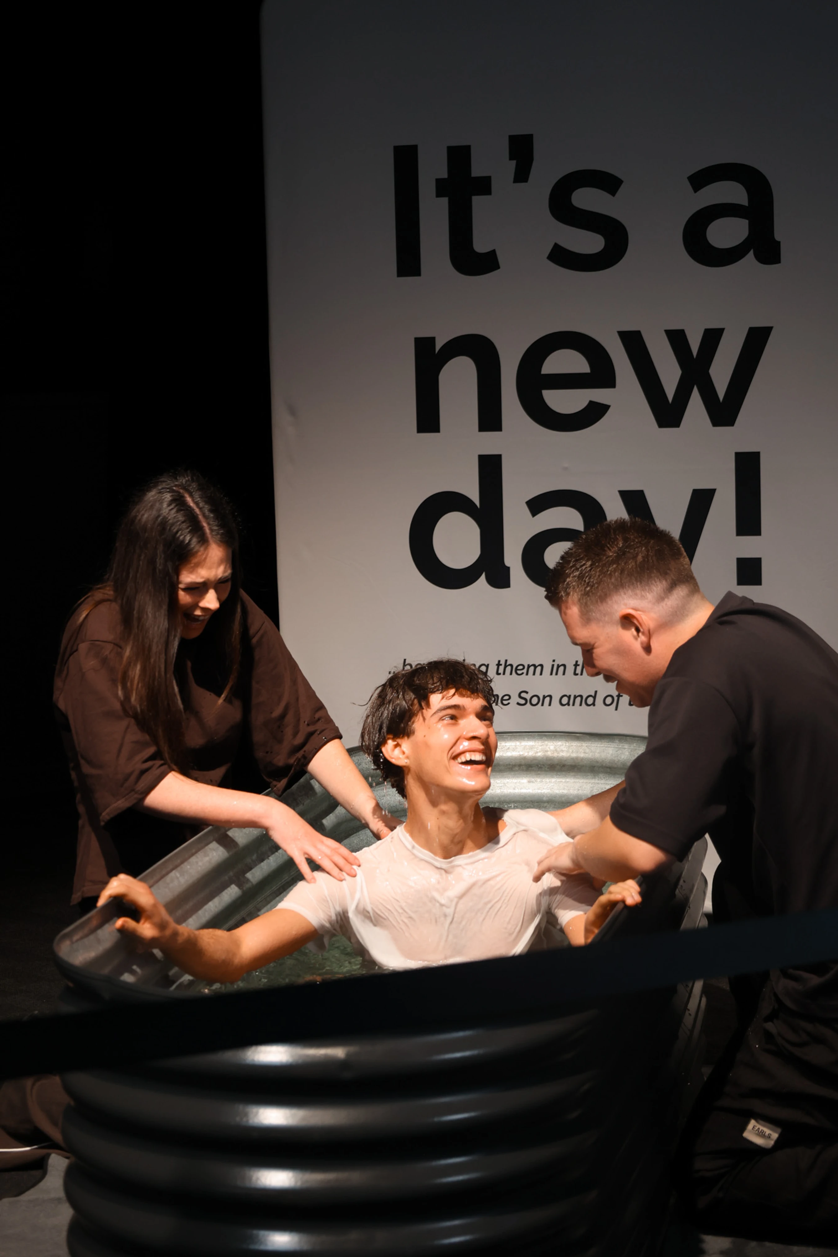 A smiling young man in a wet white shirt is supported by a man and a woman as he is baptized in a large tub. A banner behind them reads, 'It's a new day!'