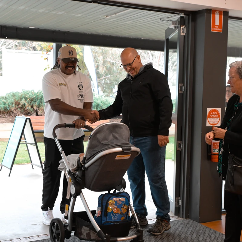 Two men smile as they shake hands in a doorway. One man has a baby in a stroller. An older woman stands nearby, looking on.