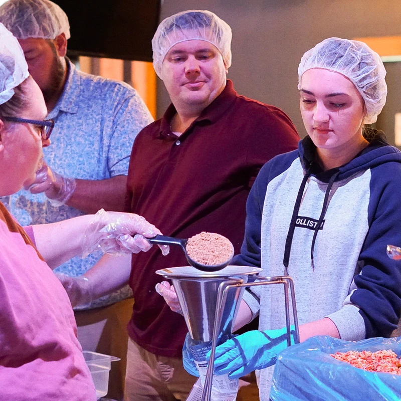 A group of volunteers wearing hairnets and gloves work at a food packing station, scooping ingredients into bags using a funnel.