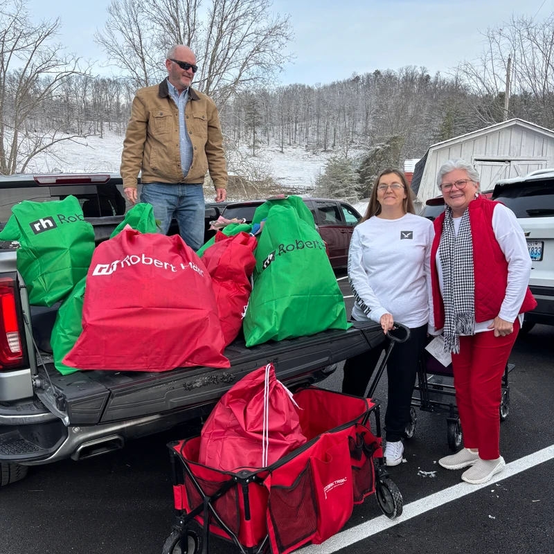 Three volunteers stand by a pickup truck filled with red and green gift bags. A man is in the truck bed and two women stand beside it on a snowy day.