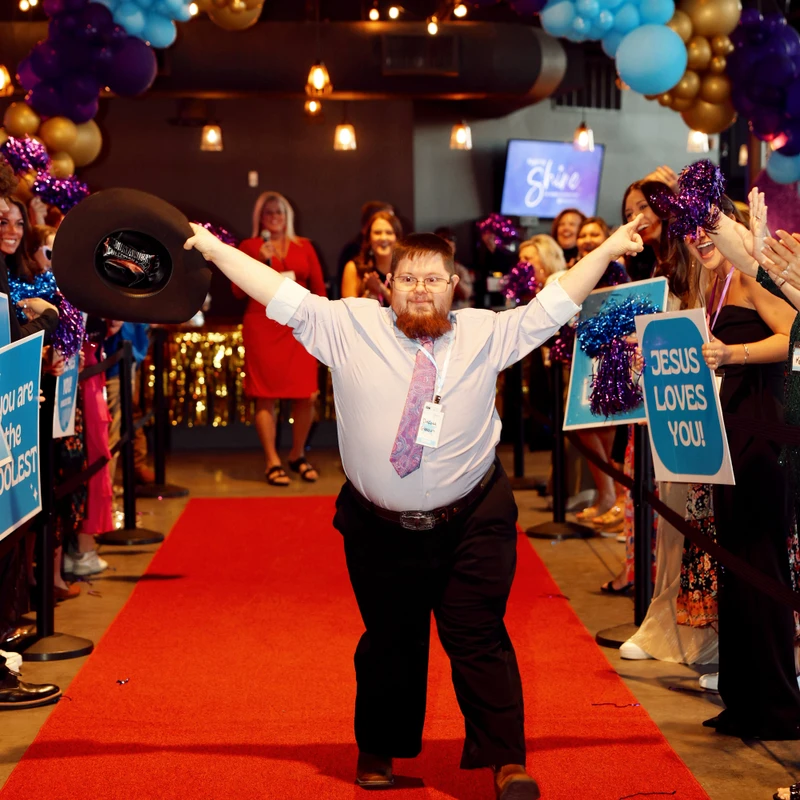 A man with a beard and glasses walks a red carpet with arms open. A crowd cheers, holding signs and pom-poms under festive balloon arches.