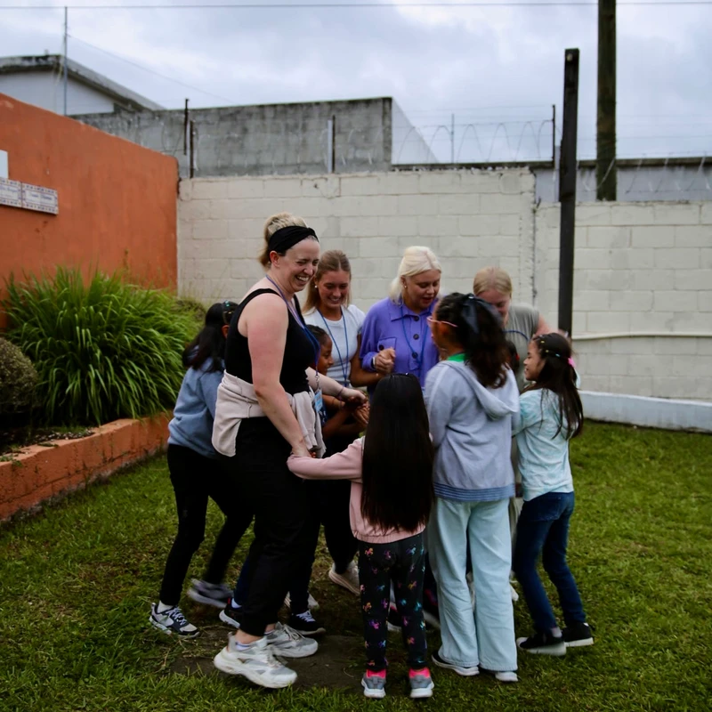 A group of women and young girls hold hands and smile while gathered in a circle on a grassy lawn in an enclosed yard.