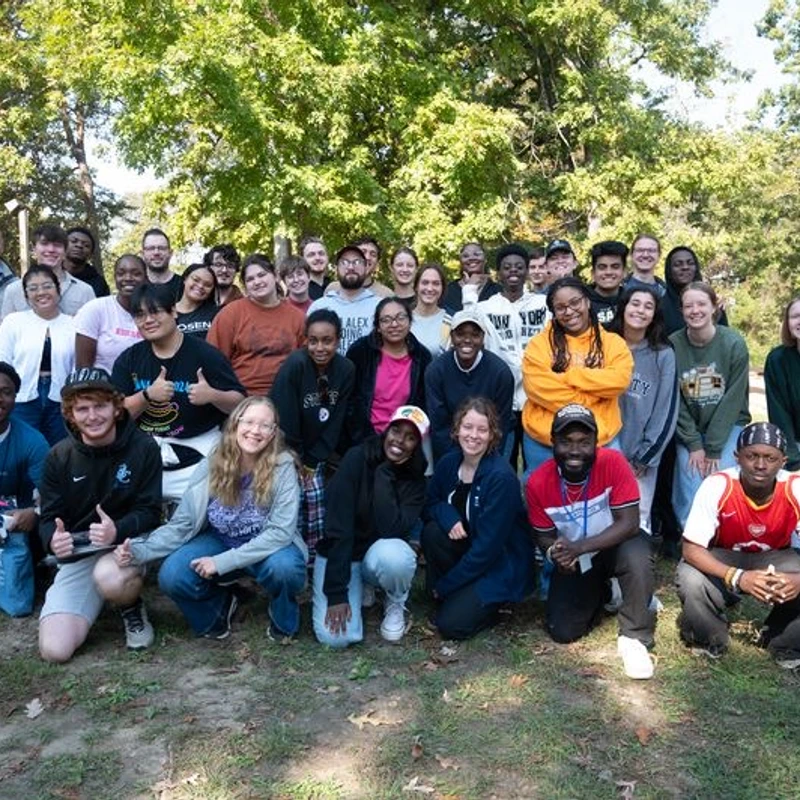 A large, diverse group of mostly young people smile for a group photo. They are gathered in a grassy, wooded area on a sunny day.