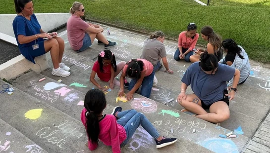 A group of children and women are gathered on wide concrete stairs, drawing colorful pictures and writing their names with sidewalk chalk in the afternoon.