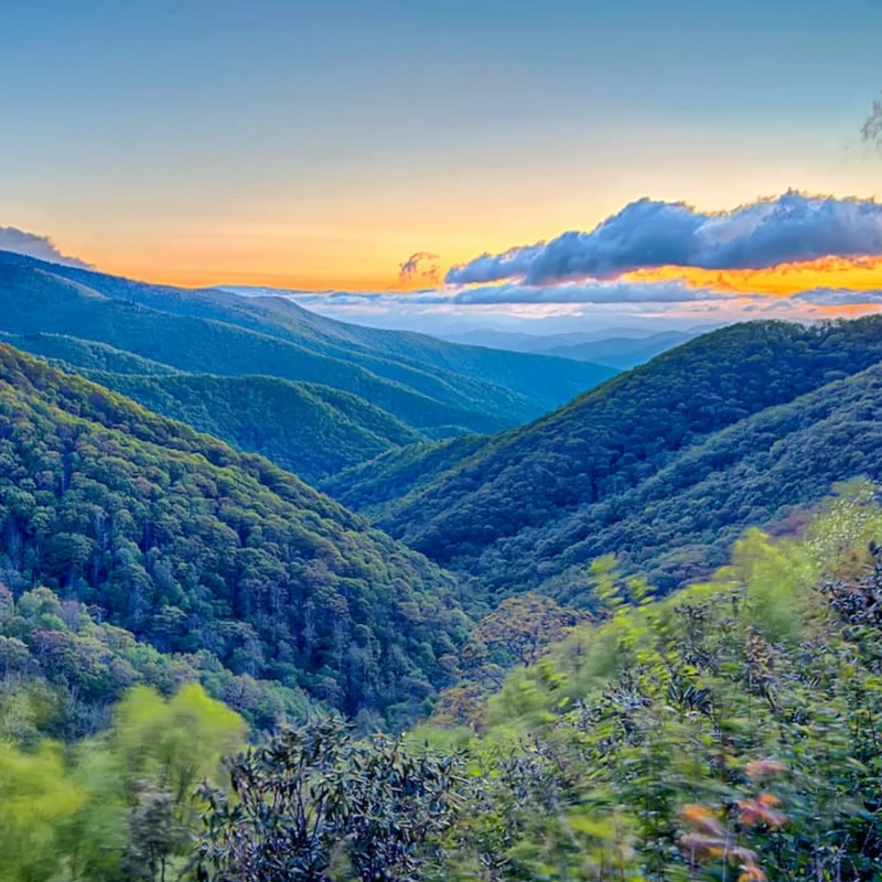 A scenic view of rolling green mountains forming a deep valley. A golden sunset with clouds illuminates the horizon above the distant peaks.