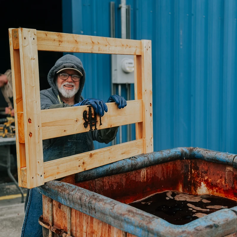 An older man in a hoodie and overalls smiles while dipping a wooden frame with a branded logo into a large rusty vat of dark liquid in a workshop.