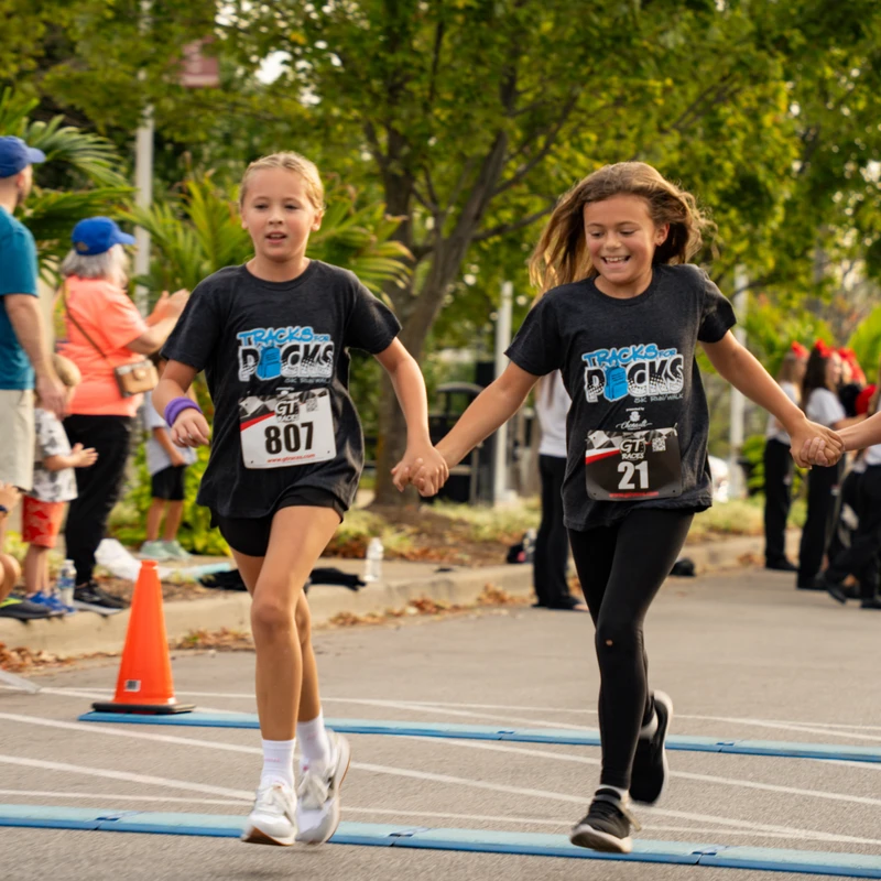 Three young girls in matching t-shirts run joyfully, holding hands as they cross blue timing mats on the street during a community race.