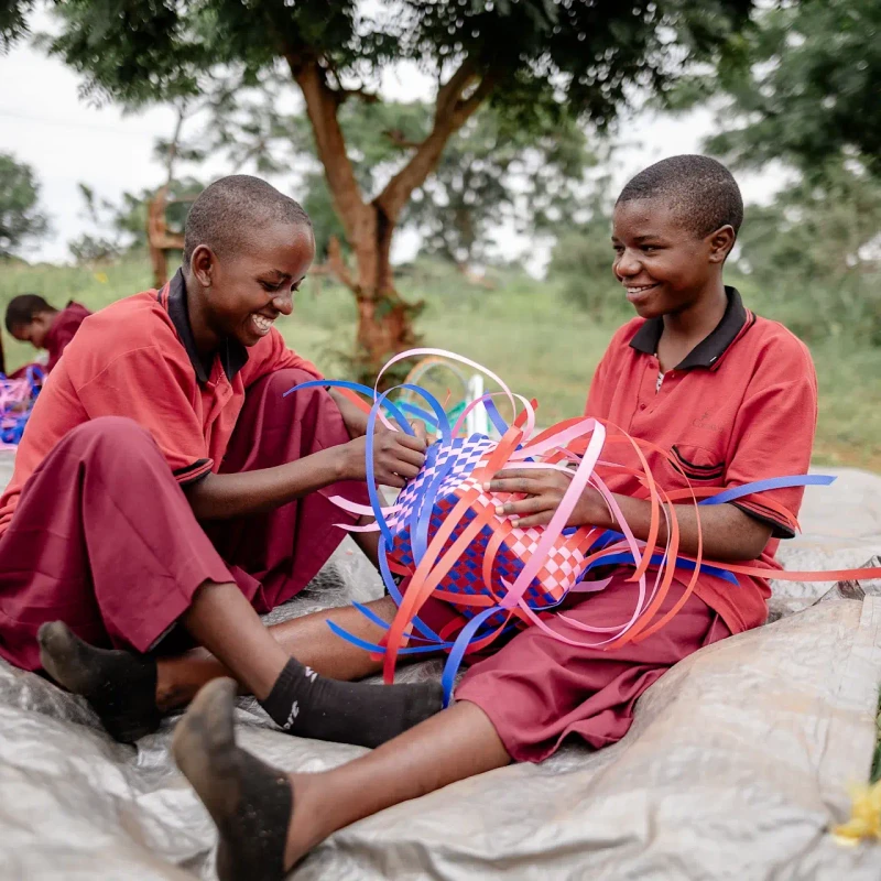 Two young students in red uniforms sit on a tarp outside, laughing and smiling as they weave a basket together from colorful blue, pink, and red ribbons.