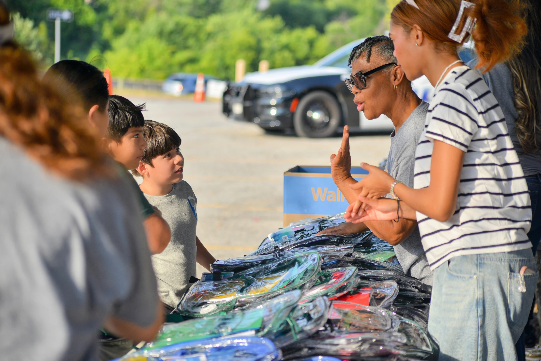 An adult volunteer in sunglasses talks to a group of children and a teen at an outdoor school supply giveaway event in a parking lot.