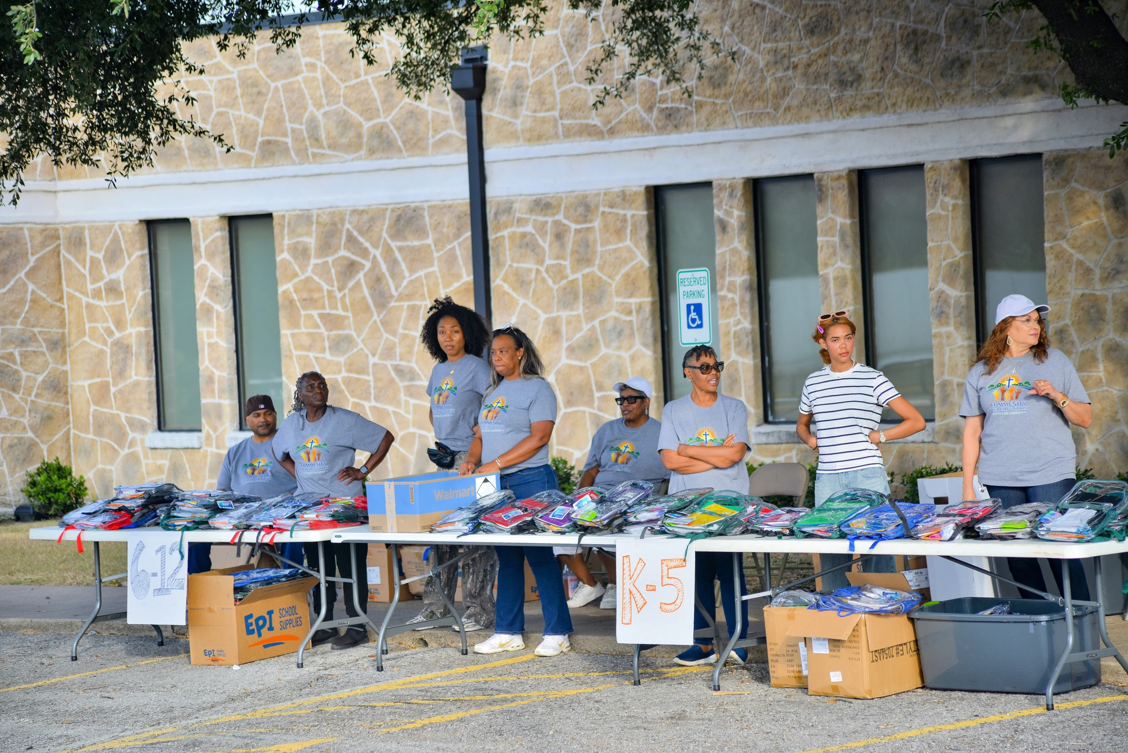A diverse group of volunteers stand behind long tables filled with packaged school supplies during an outdoor giveaway event in a parking lot.