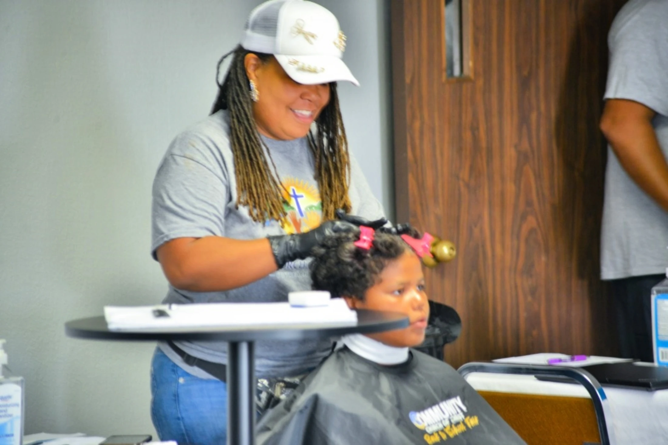 A smiling woman with braids, wearing a white cap and gloves, styles a young child's hair while the child sits patiently in a black cape.